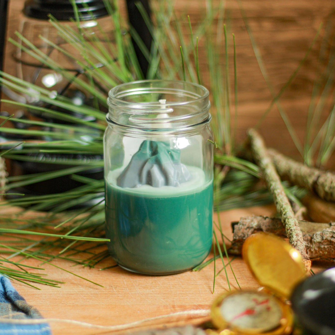 Jar with green candle on a wooden surface with plants and lantern in the background